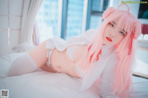 A woman with long pink hair sitting on a bed.